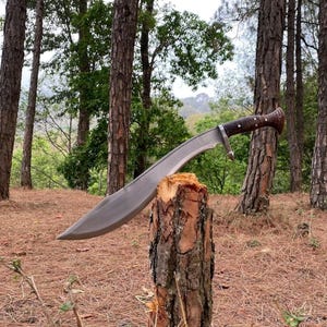 May include: A large, curved blade knife with a wooden handle and a silver-colored blade. The knife is resting on a tree stump in a forest setting, surrounded by tall trees and fallen pine needles.