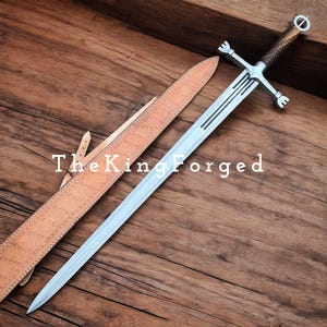 May include: A long, silver sword with a brown leather-wrapped handle and a crossguard. The blade has three dark lines etched near the center. A tan leather sheath lies beside the sword. The text "The King Forged" is visible.
