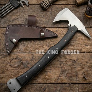 Puede incluir: Un hacha de acero forjado con mango negro y funda de cuero. El hacha tiene una hoja plateada y el texto "THE KING FORGED" es visible. El hacha est&aacute; sobre una superficie de madera.