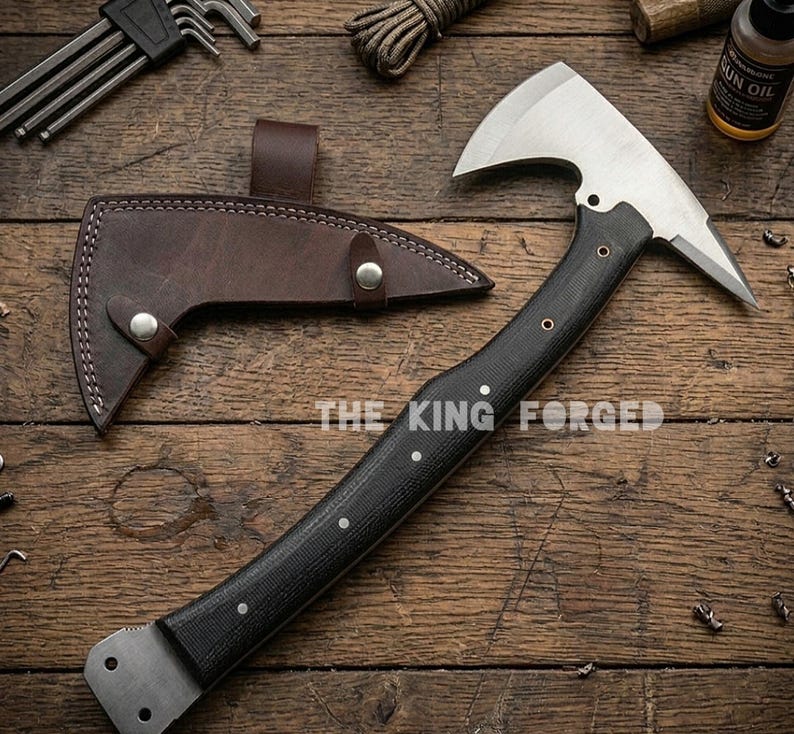 Puede incluir: Un hacha de acero forjado con mango negro y funda de cuero. El hacha tiene una hoja plateada y el texto "THE KING FORGED" es visible. El hacha est&aacute; sobre una superficie de madera.