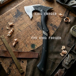 May include: A handcrafted axe with a black handle and a silver blade. The axe features the text "THE KING FORGED". The handle has two silver stripes. The axe is on a wooden surface.