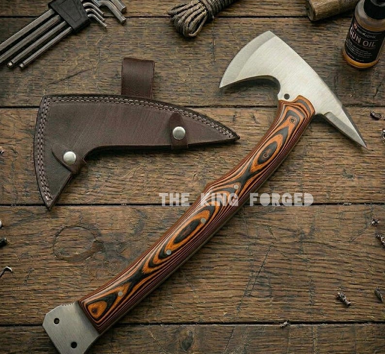 Puede incluir: Un hacha artesanal con una hoja de acero inoxidable y un mango de madera con un patr&oacute;n de vetas &uacute;nico. El hacha se muestra con una funda de cuero marr&oacute;n. El texto "THE KING FORGED" es visible en el mango.
