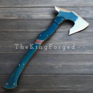 Könnte beinhalten: Eine handgefertigte Axt mit polierter Stahlklinge und gebogenem Griff. Der Griff ist tief türkisblau mit einem braunen Akzent und weißen Punktdetails. Der Text "The King Forged" ist auf dem Griff aufgedruckt.