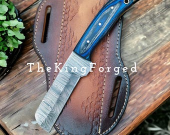 May include: A Damascus steel knife with a blue and black wooden handle, on a brown leather sheath. The knife blade has a distinctive pattern, and the sheath has embossed details. The text "The King Forged" is visible.
