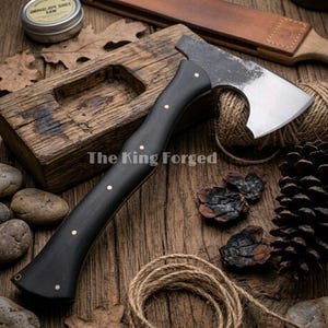 Könnte beinhalten: Eine handgefertigte Axt mit schwarzem Griff und silberner Klinge. Der Griff ist mit Messingnieten verziert. Die Axt wird auf einer Holzoberfläche mit dem Text "The King Forged" präsentiert.