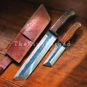 May include: Two forged steel knives with wooden handles and a brown leather sheath. The larger knife has a long, rectangular blade, while the smaller one has a shorter, angled blade. The sheath is stitched with a diamond pattern. The text "The King Forged" is visible.