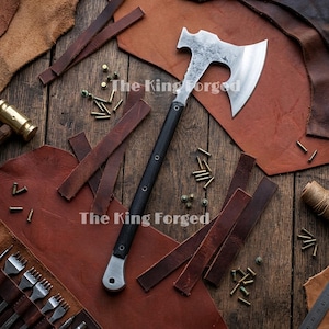 May include: A hand-forged steel axe with a black handle and a silver pommel, featuring a textured metal head with a curved blade and a hammer poll. The text The King Forged is visible on the blade. The axe is surrounded by leather scraps and metal rivets.