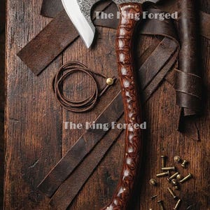 May include: A hand axe with a textured, dark metal head and a polished, contoured dark wood handle. The text 'The King Forged' is visible on the blade and handle area. Brown leather straps, a coiled leather cord, and small metal rivets are present.