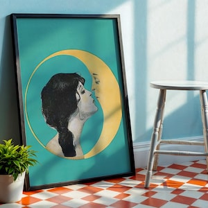 Op de afbeelding: Ingelijste kunstdruk met een vrouw en een halve maan in een turquoise cirkel. Het profiel van de vrouw is in zwart-wit, met de maan in geel. Het kunstwerk staat in een zwarte lijst, leunend tegen een muur. Een kleine potplant en een witte kruk staan op de voorgrond.