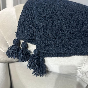 Puede incluir: Una manta texturizada azul marino con tres borlas a juego. La manta está colocada sobre una silla tapizada de color claro. La textura de la manta parece suave y afelpada, adecuada para la decoración del hogar.