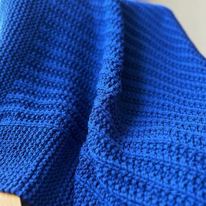 May include: A close-up of a folded, knitted blanket in a vibrant shade of blue. The blanket's texture is visible, with a combination of ribbed and textured stitch patterns. The blanket is folded over a wooden surface.