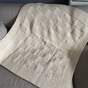 May include: A cream-colored knitted blanket with a woven pattern, draped over a gray sofa. The blanket features a textured design with a border, offering a cozy and decorative element for home decor.