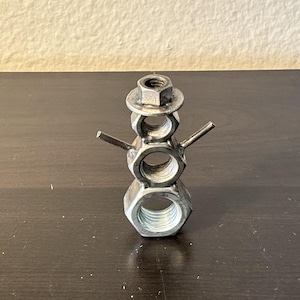 May include: A metal snowman sculpture crafted from nuts and bolts. The snowman features a hexagonal nut base, a body of stacked nuts, and a nut hat. Small metal rods serve as arms, creating a unique industrial art piece.