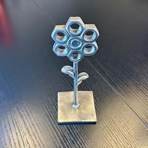May include: A handcrafted metal flower sculpture. The flower's petals are made from hexagonal nuts, arranged around a central nut. The stem and leaves are also metal, and the sculpture is mounted on a square base. The piece has a silver finish.
