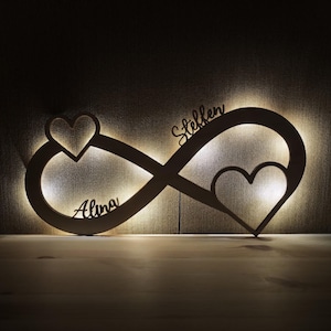 Könnte beinhalten: Ein hölzernes Unendlichkeitszeichen mit zwei Herz-Ausschnitten und den Namen "Alina" und "Stellen" in das Design geätzt. Das Symbol wird von einem warmen, weichen Licht beleuchtet, das eine romantische Atmosphäre schafft. Das Stück ist für die Wandmontage konzipiert.