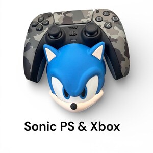 May include: A camouflage-patterned video game controller sits above a blue Sonic the Hedgehog character head. The text "Sonic PS & Xbox" is displayed below the objects. The controller has black joysticks and buttons.
