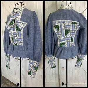 Puede incluir: Una blusa de estilo denim con detalles de patchwork. La blusa azul presenta un cuello alto, mangas largas y cintura peplum. Los detalles de patchwork incluyen patrones florales verdes, azules y blancos, con ribete de encaje.