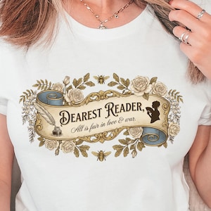 May include: White t-shirt with a banner design reading "Dearest Reader, All is fair in love & war." The banner is framed by floral and bee motifs. The shirt is worn with blue jeans.