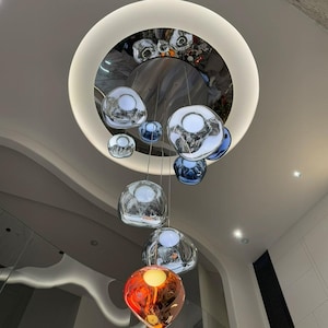Puede incluir: Una lámpara de araña con múltiples colgantes que presenta un soporte de techo circular grande con acabado cromado reflectante y luz de anillo LED integrada. Varios globos de vidrio de formas orgánicas irregulares cuelgan en tonos plata, azul y naranja.