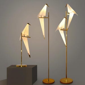 Puede incluir: Un conjunto de lámparas modernas con forma de pájaro. Las lámparas presentan alas blancas con forma de papel y marcos de metal dorado. Las lámparas están iluminadas, creando un brillo cálido. Una es una lámpara de mesa, las otras son lámparas de pie.