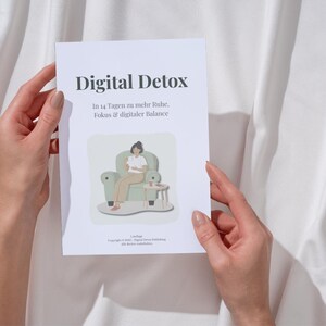Könnte beinhalten: Ein weißes Buch mit dem Titel "Digital Detox" wird von zwei Händen vor einem weißen Stoffhintergrund gehalten. Das Cover des Buches zeigt Text und eine Illustration einer Person in einem Stuhl. Der Untertitel des Buches lautet "In 14 Tagen zu mehr Ruhe, Fokus & digitaler Balance."