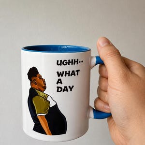 May include: A white ceramic mug with a blue interior and handle. The mug features a graphic illustration of a person looking upward with the text "UGH... WHAT A DAY" printed in black, bold, sans-serif capital letters on the front surface.