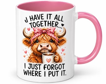 May include: A white ceramic mug with a pink interior and handle, featuring a graphic of a brown highland cow wearing a pink polka-dot bow. The text reads: I HAVE IT ALL TOGETHER... I JUST FORGOT WHERE I PUT IT. Small pink hearts surround the cow.