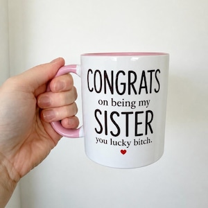 Peut inclure: Un mug en céramique blanche avec un intérieur et une anse rose clair. L'avant présente un texte noir qui dit CONGRATS on being my SISTER you lucky bitch. Un petit cœur rouge est imprimé sous le texte. Le mug a une forme cylindrique.
