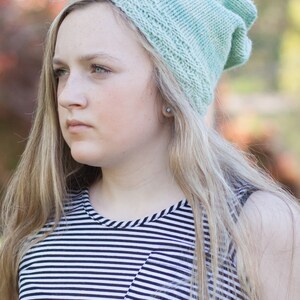 May include: A young person with long blonde hair wearing a light green knitted beanie and a black and white striped top.