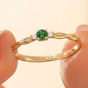 May include: A gold ring featuring a central round green gemstone flanked by two smaller clear stones. The band has a twisted design and is engraved with the text VOQ on the inner surface. The ring is held between two fingers.