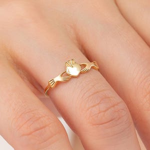 May include: A gold Claddagh ring. The ring features a heart held by two hands, topped with a crown. The ring is worn on a finger, showcasing its intricate design and polished finish. The ring is a symbol of love, loyalty, and friendship.