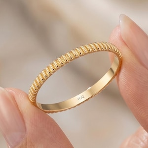 Könnte beinhalten: Ein goldener Ring mit einem strukturierten, geriffelten Design. Der Ring wird von einer Person gehalten. Der Ring ist ein einfaches Band mit einem sich wiederholenden Muster aus vertikalen Linien. Der Ring hat eine helle, glänzende Goldfarbe.