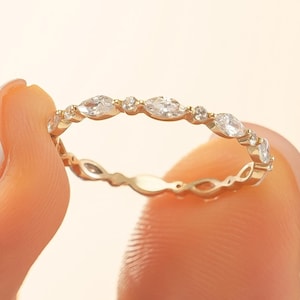 May include: A delicate gold ring with a repeating pattern of marquise-cut and round clear gemstones. The ring is held between two fingers against a soft, neutral background. The ring's band has a subtle, openwork design.