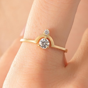 May include: A gold ring with a crown design, featuring a large, round, clear gemstone at the centre and a smaller, teardrop-shaped gemstone above. The ring is on a finger, showcasing its delicate details and elegant style.