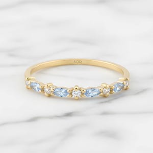 May include: A gold ring with a delicate design, set with alternating light blue and white gemstones. The ring features marquise-cut blue stones and round white stones. The ring is displayed on a white marble surface.