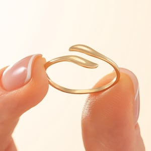 May include: A gold-colored ring with a unique, open design. The ring features a slender band that curves around the finger, with two elongated, teardrop-shaped ends that overlap slightly. The ring is held between two fingers.