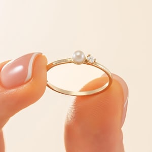 May include: A delicate gold ring featuring a small, round pearl and a tiny, sparkling gemstone. The ring has a thin band and a minimalist design. The ring is held up against a soft, neutral background.