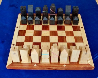 May include: A wooden chess set with light and dark brown pieces on a wooden board with a light and dark brown checkerboard pattern. The pieces are shaped like houses.