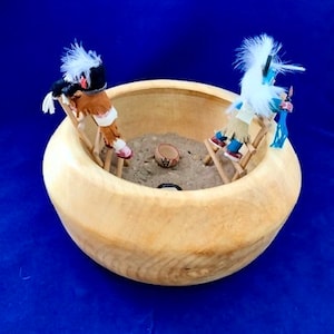 May include: Two Native American dolls, one wearing a blue and white outfit and the other wearing a red and white outfit, are sitting on wooden ladders inside a wooden bowl. The bowl is filled with sand and a small clay pot.