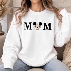May include: White sweatshirt with the word "MOM" in black block letters, with a Mickey Mouse head replacing the "O". The sweatshirt has long sleeves and a crew neck. The overall theme is Disney-inspired apparel.