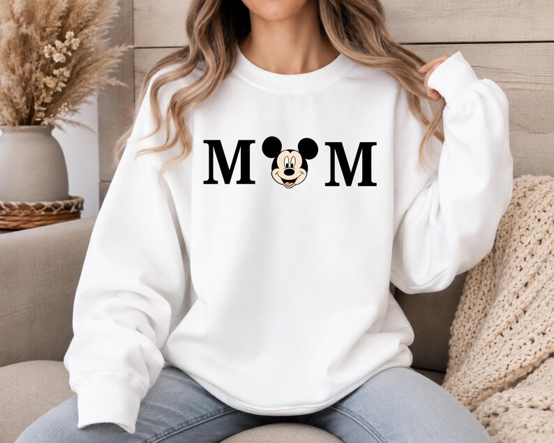 May include: White sweatshirt with the word "MOM" in black block letters, with a Mickey Mouse head replacing the "O". The sweatshirt has long sleeves and a crew neck. The overall theme is Disney-inspired apparel.