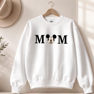 May include: White sweatshirt with the word "MOM" in black block letters, with a Mickey Mouse head replacing the "O". The sweatshirt is hanging on a wooden hanger.