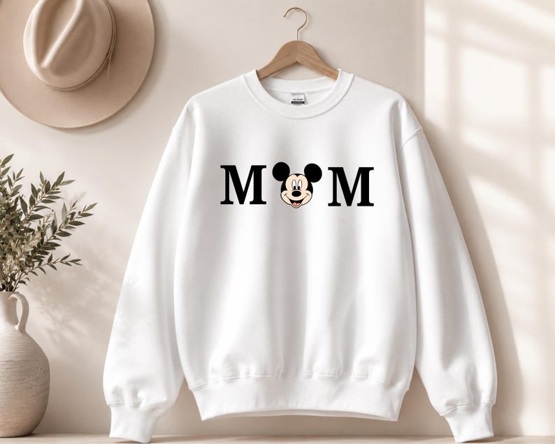 May include: White sweatshirt with the word "MOM" in black block letters, with a Mickey Mouse head replacing the "O". The sweatshirt is hanging on a wooden hanger.