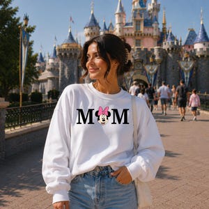 May include: A white sweatshirt with the word "MOM" in black letters, featuring a Minnie Mouse graphic with a pink bow. The sweatshirt is worn by a person in front of a castle.