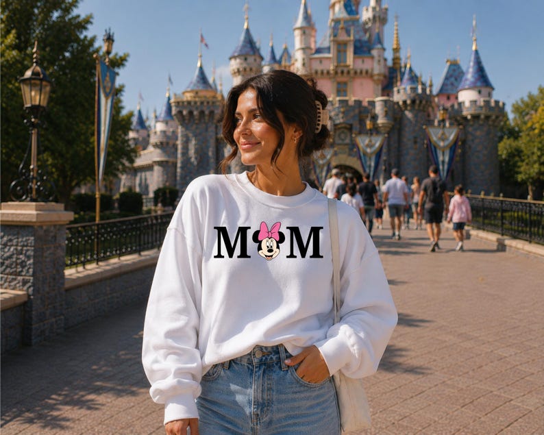 May include: A white sweatshirt with the word "MOM" in black letters, featuring a Minnie Mouse graphic with a pink bow. The sweatshirt is worn by a person in front of a castle.