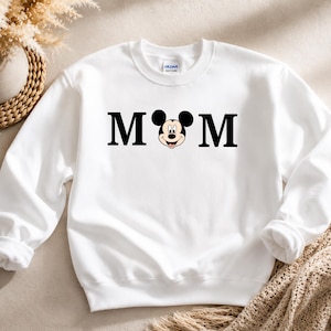 May include: White sweatshirt with the word "MOM" in black block letters, with Mickey Mouse's face replacing the "O". The sweatshirt has a crew neck and long sleeves. The brand name "Gildan" is visible on the tag.