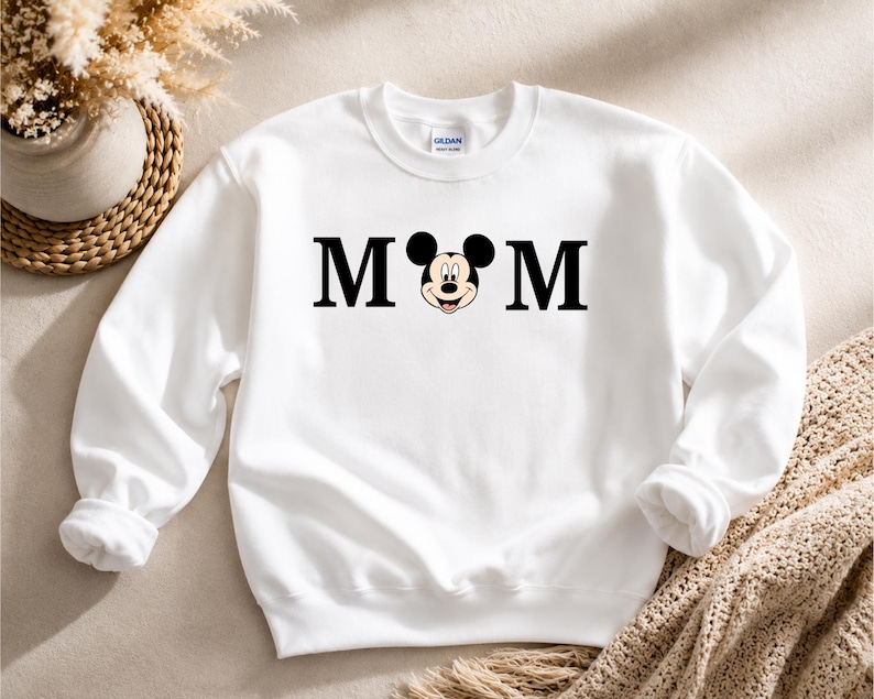 May include: White sweatshirt with the word "MOM" in black block letters, with Mickey Mouse's face replacing the "O". The sweatshirt has a crew neck and long sleeves. The brand name "Gildan" is visible on the tag.