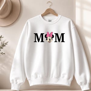 May include: White sweatshirt with the word "MOM" in black block letters. A Minnie Mouse graphic with a pink bow replaces the "O". The sweatshirt is hanging on a wooden hanger.