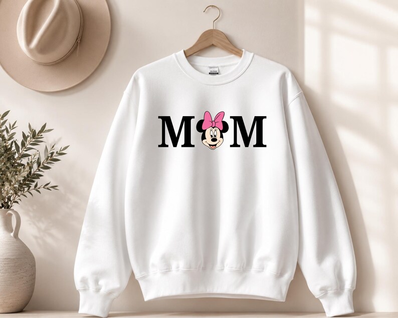 May include: White sweatshirt with the word "MOM" in black block letters. A Minnie Mouse graphic with a pink bow replaces the "O". The sweatshirt is hanging on a wooden hanger.