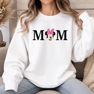 May include: White sweatshirt with the word "MOM" in black block letters. The "O" is replaced with a Minnie Mouse graphic wearing a pink bow. The sweatshirt has long sleeves and a crew neck.